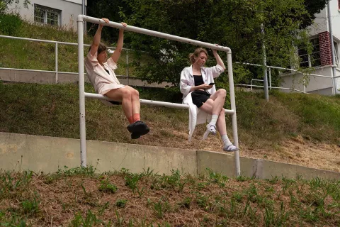 Zwei Personen sitzen nebeneinander auf einem weißen Geländer, das parallel zu einem Hang verläuft. Eine Person hängt mit beiden Armen an der oberen Querstange und blickt konzentriert zur Seite, die Beine angewinkelt über der unteren Strebe. Die andere Person sitzt entspannt mit überschlagenen Beinen, eine Hand am Geländer, die andere hält ein Smartphone. Im Hintergrund: Hang mit Wiese, Sträuchern und Wohngebäuden mit roten Fensterläden.