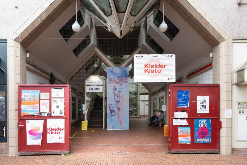 Das Bild zeigt den Eingang der ehemaligen Schwabenbräu-Passage in Bad Cannstatt mit moderner Architektur, Plakaten an den roten Toren und einer Sitzbank.