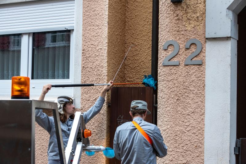 Das Bild zeigt zwei Personen in grauer Arbeitskleidung, die mit Reinigungsgeräten eine Hausfassade mit der Nummer 22 reinigen. Eine Person benutzt eine blaue Bürste an einem langen Stab, um schwer erreichbare Stellen zu säubern.