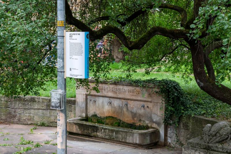 Das Bild zeigt ein Hinweisschild über eine Soundinstallation, das an einer Straßenplanke vor einem Brunnen befestigt ist.