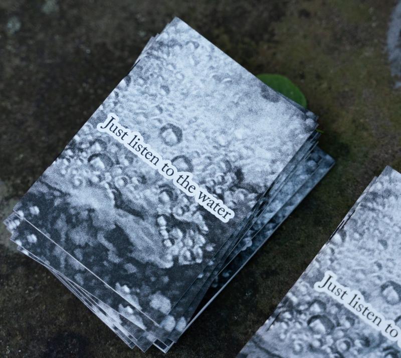 Das Bild zeigt einen Stapel schwarz-weißer Flyer mit der Aufschrift „Just listen to the water“. Die Struktur des Wassers ist als Hintergrundmotiv auf den Flyern gedruckt.