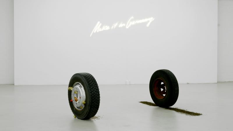 Das Bild zeigt eine minimalistische Kunstausstellung mit zwei stehenden Autoreifen, aus denen Erde und Lebensmittel herausquellen, vor einer weißen Wand mit der leuchtenden Schrift „Make it in Germany“.