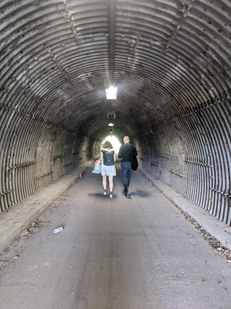 Zwei Personen durchqueren einen schmalen, geriffelten Fußgängertunnel – ein alltäglicher Raum, der durch O.J.A.I.s Intervention zum Ort spekulativer Infrastrukturpoesie wird.