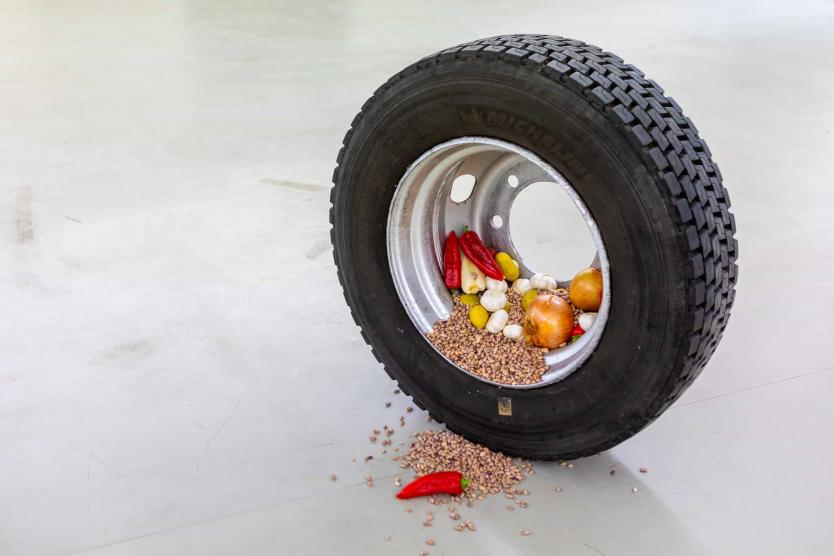 The image shows a car tire lying on the ground, filled with various foods such as onions, garlic, chili peppers, and grains, some of which have fallen onto the floor.
