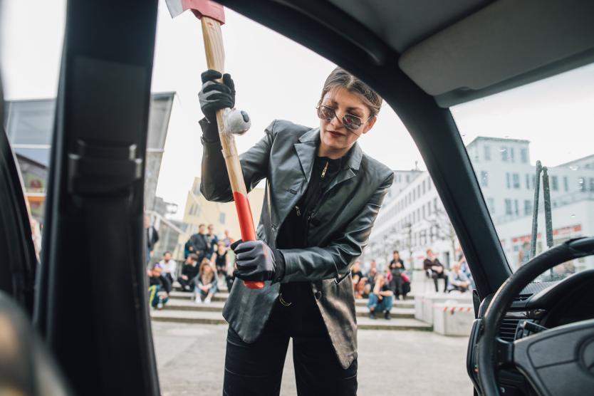 Das Bild zeigt eine Person in schwarzer Lederkleidung, die mit ernstem Gesichtsausdruck und erhobener Axt durch das geöffnete Autofenster schaut, während im Hintergrund Menschen auf Stufen sitzen und zusehen.