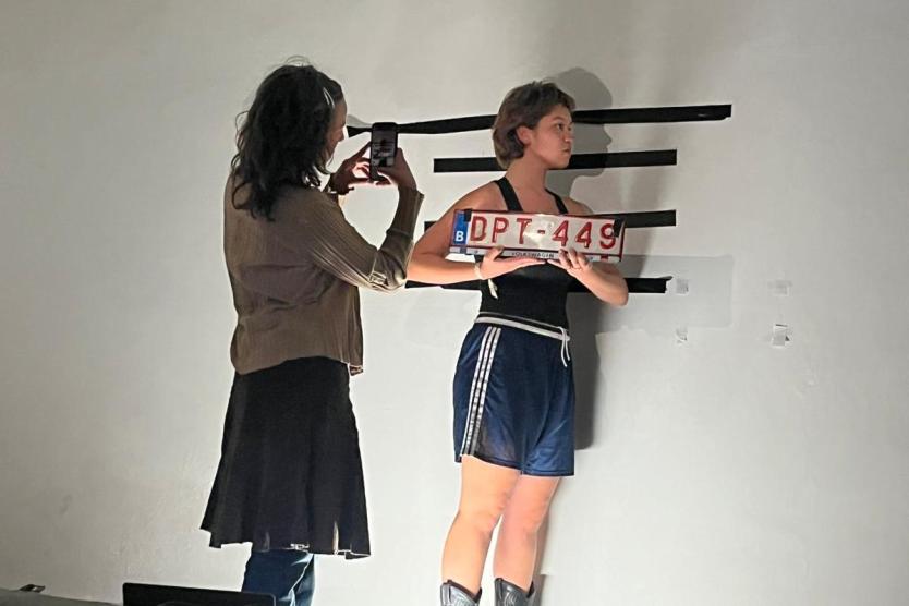 The image shows a person wearing shorts and cowboy boots standing in front of a white wall, holding a license plate with the inscription "DPT-449," while another person is taking a photo of them with a smartphone.