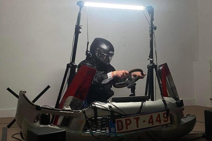 The image shows a person wearing a motorcycle helmet and leather jacket, sitting on an improvised driving simulator made from car parts, with the license pThe image shows a person wearing a motorcycle helmet and leather jacket, sitting on an improvised driving simulator made from car parts, with the license plate "DPT-449." They are surrounded by light and technical devices.