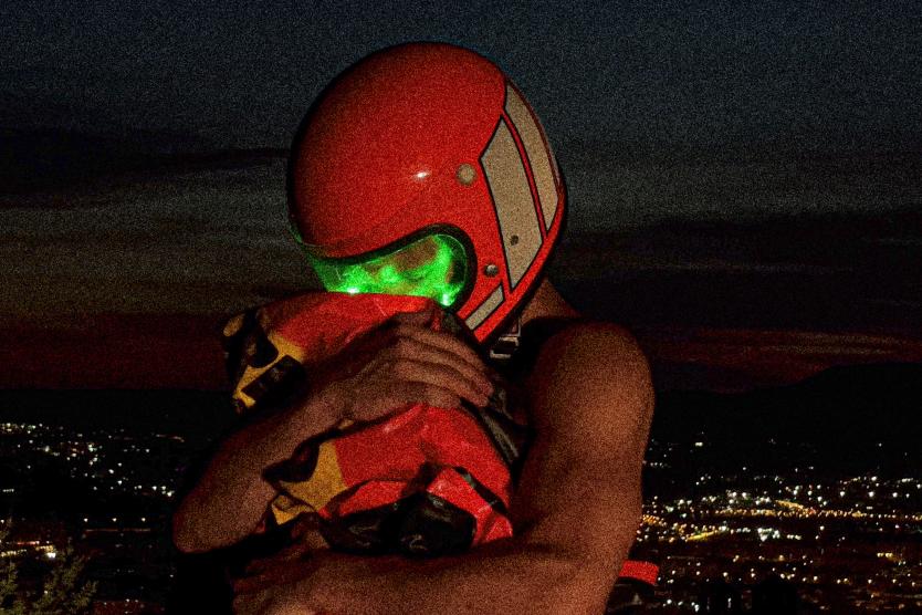 The image shows a person wearing an orange helmet and a bright green visor, tightly hugging an orange package, with a nighttime cityscape and glowing lights in the background.