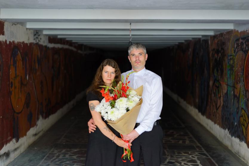 The image shows a couple or two people standing together in elegant clothing, embracing with serious expressions in a graffiti-covered, dark tunnel. They are holding a large bouquet of white and red flowers.