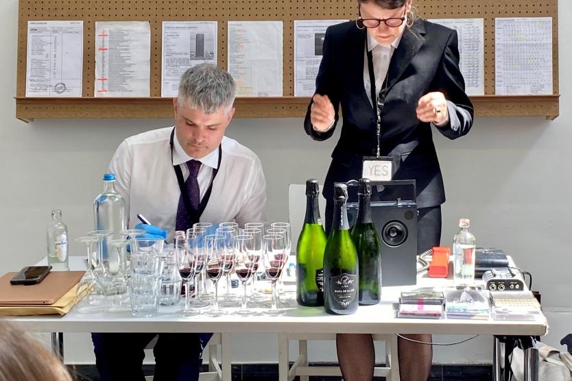 The image shows two people in formal clothing sitting at a table full of glasses, champagne bottles, and technical devices, working intently. One person is standing and gesturing, while the other is writing something down.