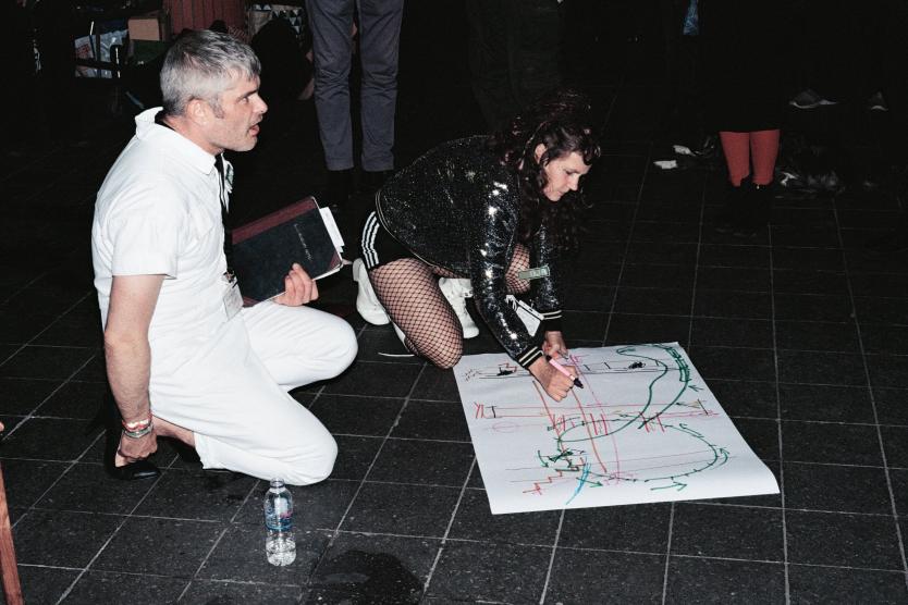 The image shows a person in a white overall kneeling, interacting with another person in a glittering outfit and fishnet stockings, while the latter is creating a large drawing with colorful lines and symbols on the floor.