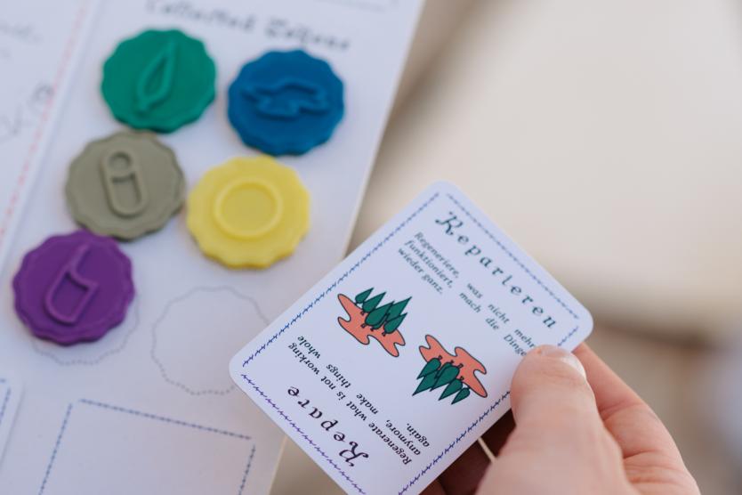 Das Bild zeigt eine Hand, die eine Spielkarte mit der Aufschrift „Repariere“ hält; daneben sind auf dem Spielbrett mehrere farbige Token angeordnet.