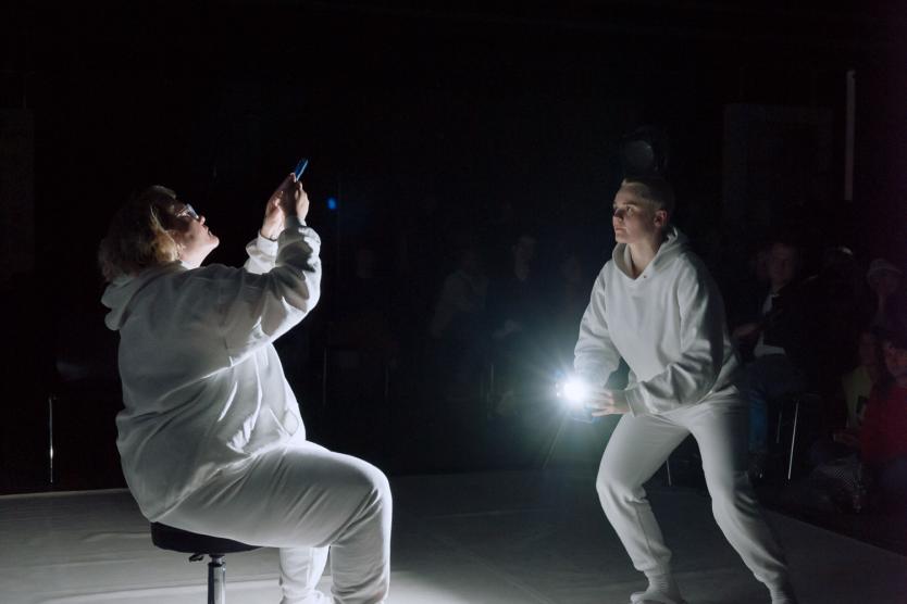 The image shows the two artists during a performance. They are wearing white full-body suits, and Julia is directing a flashlight at Carmen, who is sitting on a chair and holding her hands protectively in front of her face.