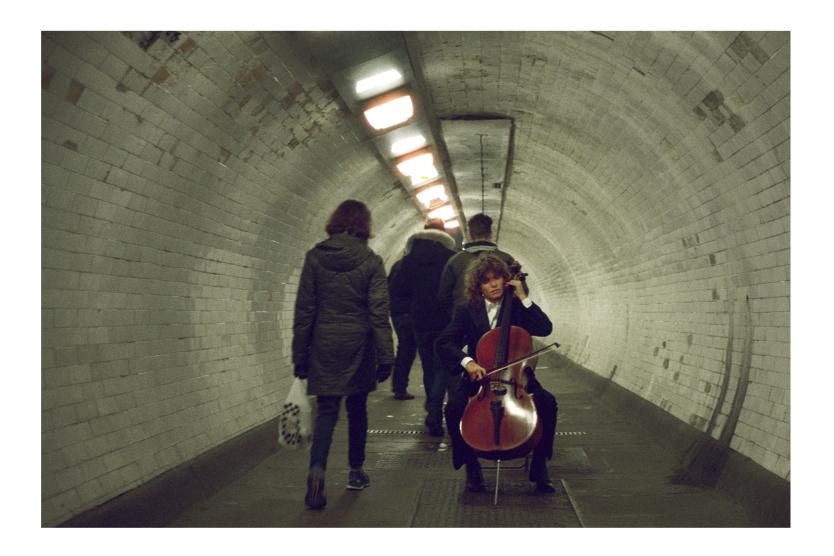 In einem gekachelten Fußgängertunnel spielt eine Person Cello, während mehrere andere Personen daran vorbeigehen. Die Musikerin sitzt auf einem Stuhl und ist konzentriert auf das Spiel. Die Szene wirkt ruhig und kontrastiert mit der Bewegung der Passantinnen im Alltag.