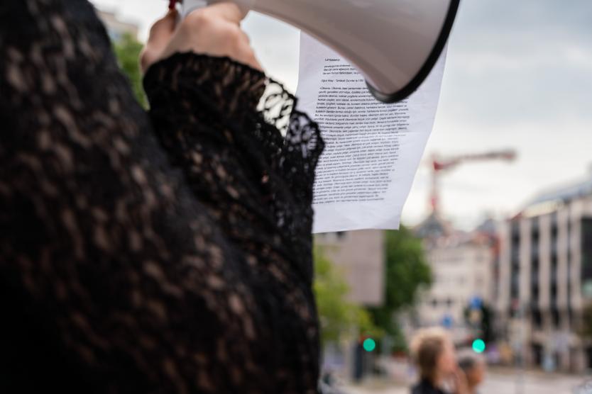 Eine Nahaufnahme zeigt ein Blatt mit türkischem Text, das an einem Megafon befestigt ist, während eine Person in schwarzer Spitze spricht und im unscharfen Hintergrund städtische Gebäude und ein Baukran sichtbar sind.