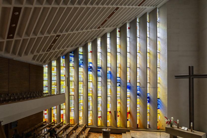 Moderne Kirche mit buntem Glasfensterband, schlichtem Altar und Kreuz. Licht fällt farbig in den Raum.