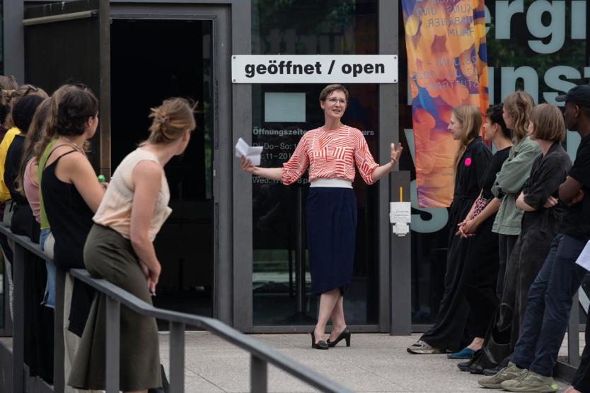 Person spricht gestikulierend vor einem Gebäude mit „geöffnet / open“-Schild, umgeben von einer zuhörenden Gruppe. Szene wirkt wie eine offizielle Begrüßung oder Einführung.