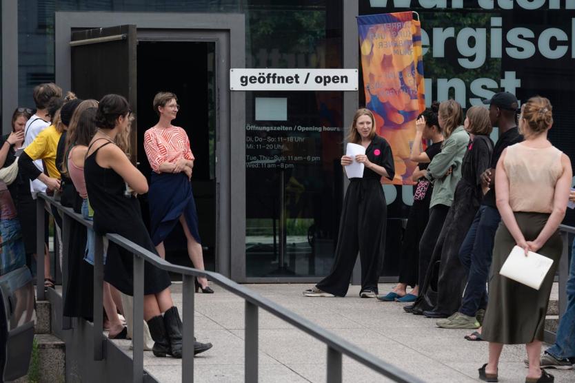 Gruppe vor dem Eingang eines Kunstinstituts, zwei Personen mit Redebeiträgen, offene Tür, Schild mit „geöffnet / open“.