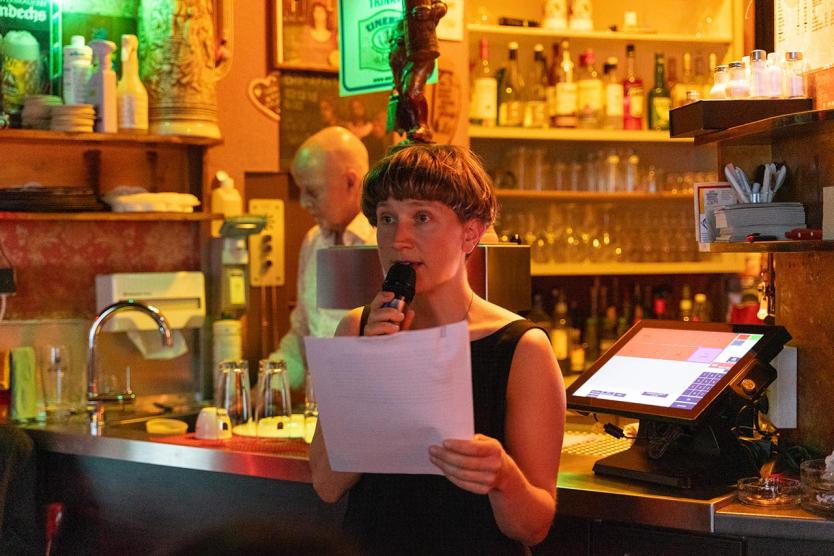 Eine Person mit liest in einer Bar von einem Blatt Papier vor. Sie spricht ins Mikrofon und hält das Blatt Papier mit Markierungen in der anderen Hand. 
