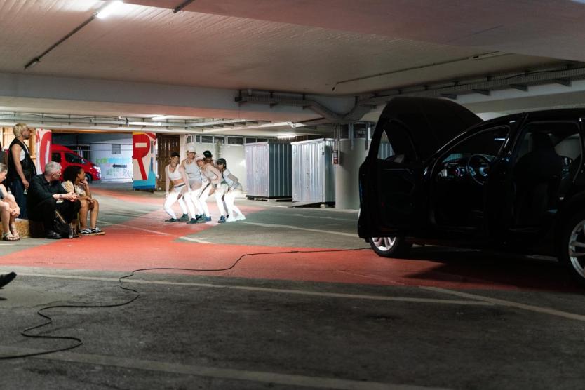 Fünf Performer*innen in weißer Kleidung posieren synchron auf einer rot markierten Fläche im Parkhaus, links sitzt Publikum auf Strohballen, rechts steht ein schwarzes Auto mit geöffneten Türen und Motorhaube.
