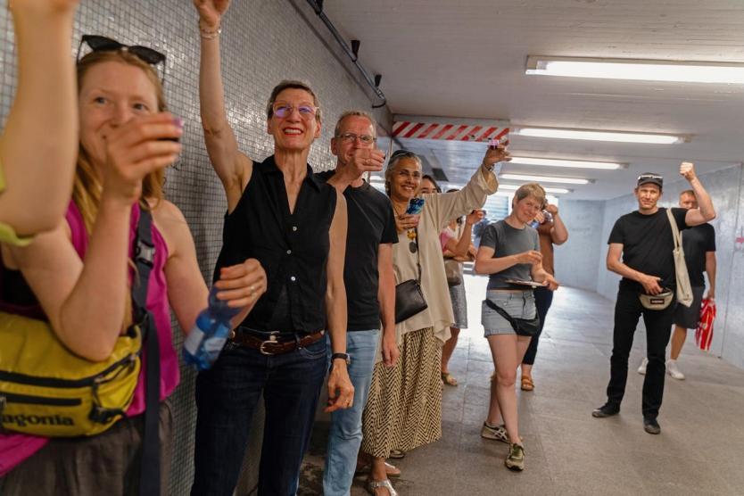 Personengruppe in einem gefliesten Tunnel hebt Becher in die Luft. Einige lächeln, andere schauen gespannt. Die Szene wirkt wie ein kollektiver Moment oder eine partizipative Geste.