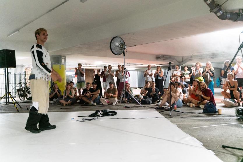 Performer auf weißer Bühne in Parkhaus, Publikum sitzt dicht gedrängt und applaudiert. Scheinwerfer und Mikrofone betonen die performative Szene.
