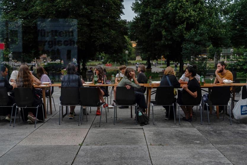 Eine Gruppe von Menschen sitzt bei einem sommerlichen Abendessen an einer langen Tafel im Freien, umgeben von Bäumen und Parklandschaft. Die Szene wirkt gesellig und entspannt.