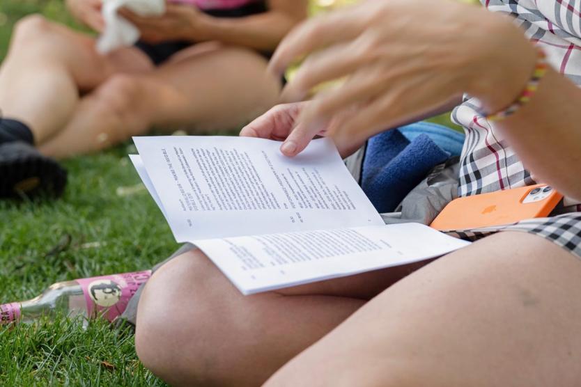 Nahaufnahme einer Person, die auf der Wiese sitzt und ein aufgeschlagenes Heft oder Buch liest. Neben ihr liegt ein orangefarbenes Smartphone, im Hintergrund eine weitere Person im Gras.