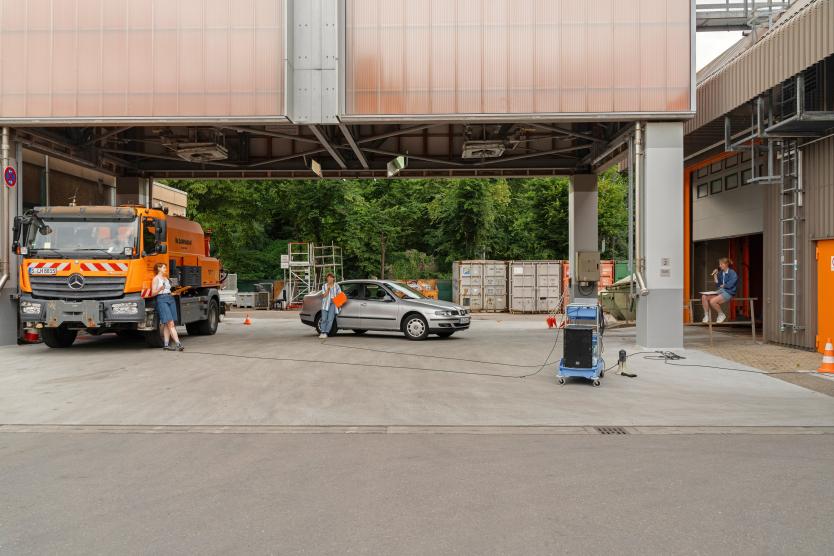 Außenbereich eines Betriebshofs mit einem orangen Lkw und einem grauen Auto, neben denen Performance-Künstlerinnen stehen.  An industrial depot area featuring an orange truck and a silver car with performance artists positioned nearby. 