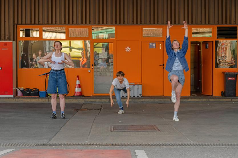 Drei Performerinnen in Alltagskleidung führen synchrone Körperbewegungen vor einer leuchtend orangen Gebäudefassade aus.  Three performers executing bodily movements outdoors in front of a bright orange building facade. 