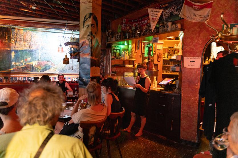 Eine Performerin steht in einer gemütlich beleuchteten Kneipe hinter der Theke und liest aus Zetteln vor, während Gäste an Tischen im Raum zuhören.  A performer stands behind the bar in a dimly lit, cozy pub, reading aloud from sheets of paper to sitting audience members.