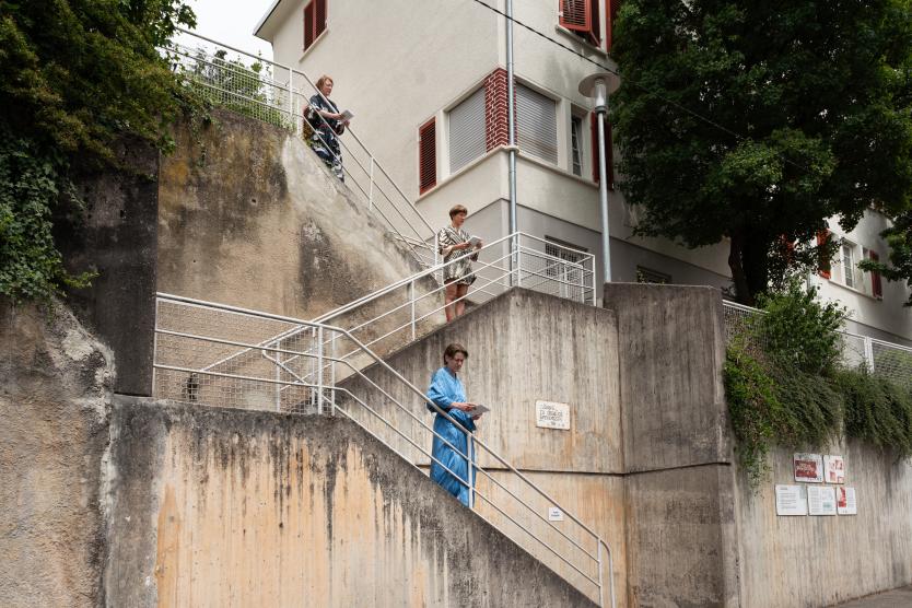 Drei Performerinnen stehen in unterschiedlichen Höhen auf den Absätzen einer betonierte Außentreppe an einem Wohngebäude und halten Blätter in den Händen.  Three performers stand at different levels on a concrete outdoor staircase next to a residential building, each holding sheets of paper.