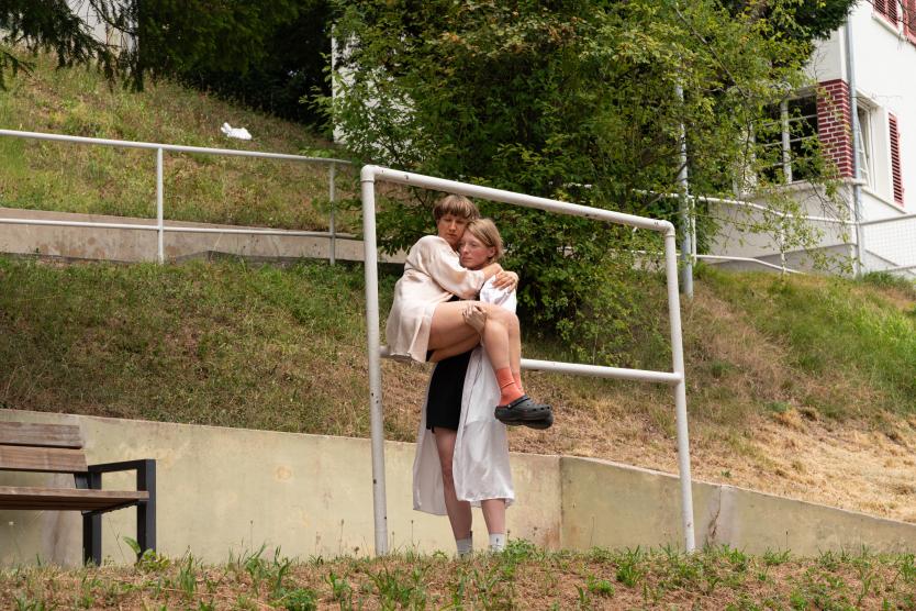 Eine Person trägt eine andere auf dem Arm im Freien vor einem Geländer an einem grasbewachsenen Hang.  One performer carries another in their arms outdoors in front of a metal railing on a grassy slope.