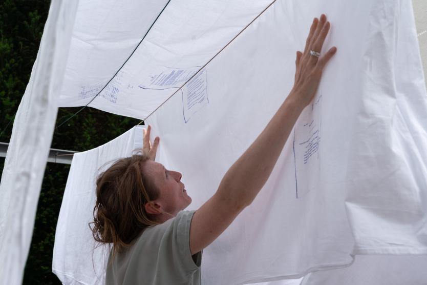 Nahaufnahme einer Frau, die weiße Bettlaken auf einer Wäscheleine aufhängt, auf denen blauer Text aufgedruckt ist.  Close-up of a woman hanging white bedsheets printed with blue text onto an outdoor clothesline.