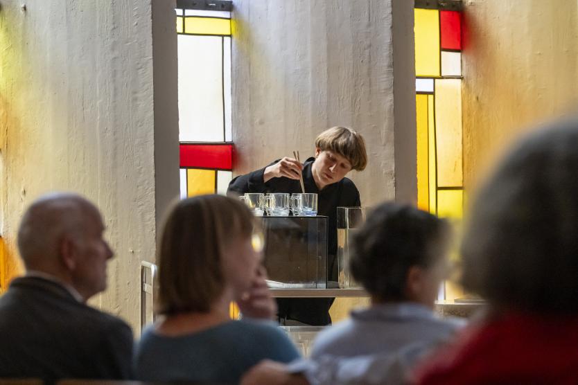 Eine Person sitzt drinnen an einem Tisch hinter einer Glaspercussion und schlägtGläser an, während das Publikum im Vordergrund zuschaut.  A performer sits indoors at a table playing water-filled glass vessels while viewed from behind members of the audience.