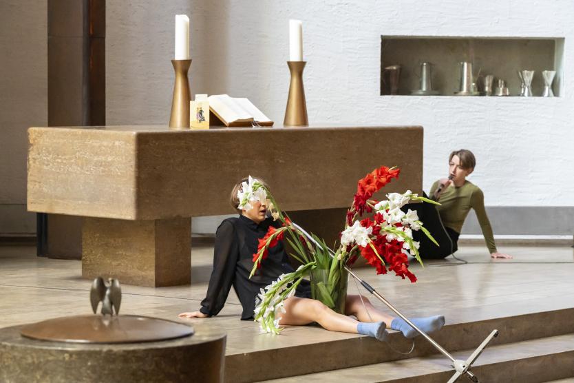 Innenansicht eines Kirchenraums: Eine Person lehnt am Altarboden neben einer großen Vase mit roten und weißen Gladiolen, während eine andere Person im Hintergrund ein Mikrofon hält.  Inside a church space, one performer reposes on the floor near a large arrangement of red and white gladioluses while another holds a microphone in the background.