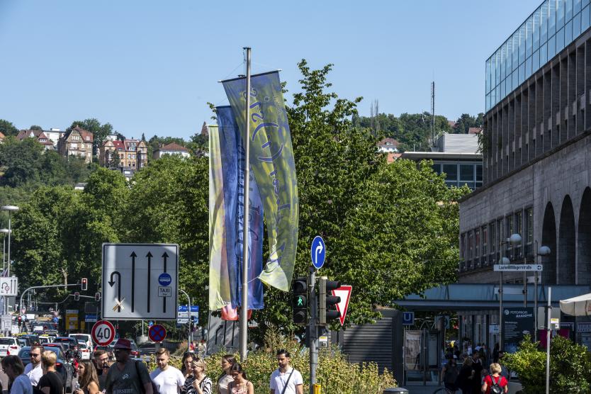 Straßenszene in einer Stadt mit bunten Fahnen im Vordergrund, Straßenschildern und Passanten, die an einer Fußgängerampel unterwegs sind.  Bustling city street scene with tall decorative banners, traffic signs, and pedestrians walking near a crosswalk.