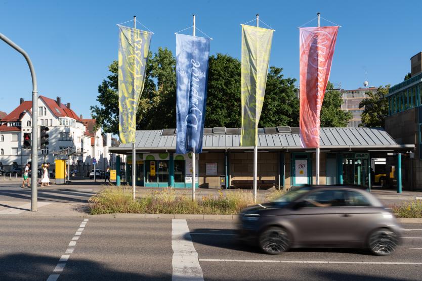 Vier lange, bunte vertikale Stoffbanner hängen an Masten vor einem flachen Gebäude an einer Straße, während ein braunes Auto im Vordergrund vorbeifährt.  Four tall, colorful fabric banners hang vertically on poles in front of a building while a blurred car drives past in the foreground.