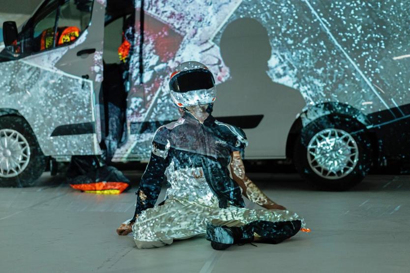 Ein Performer in Motorradkleidung und mit weiß-rotem Helm sitzt im Schneiderseitz auf dem Boden vor einem weißen Lieferwagen. Eine Projektion erzeugt ein schwarz-weißes Muster auf dem Fahrzeug, dem Helm und der Kleidung des Performers.  A performer in motorcycle gear and a white-and-red helmet sits cross-legged on the floor in front of a white van. A projection creates a black-and-white patterned visual across the vehicle, helmet, and clothing.
