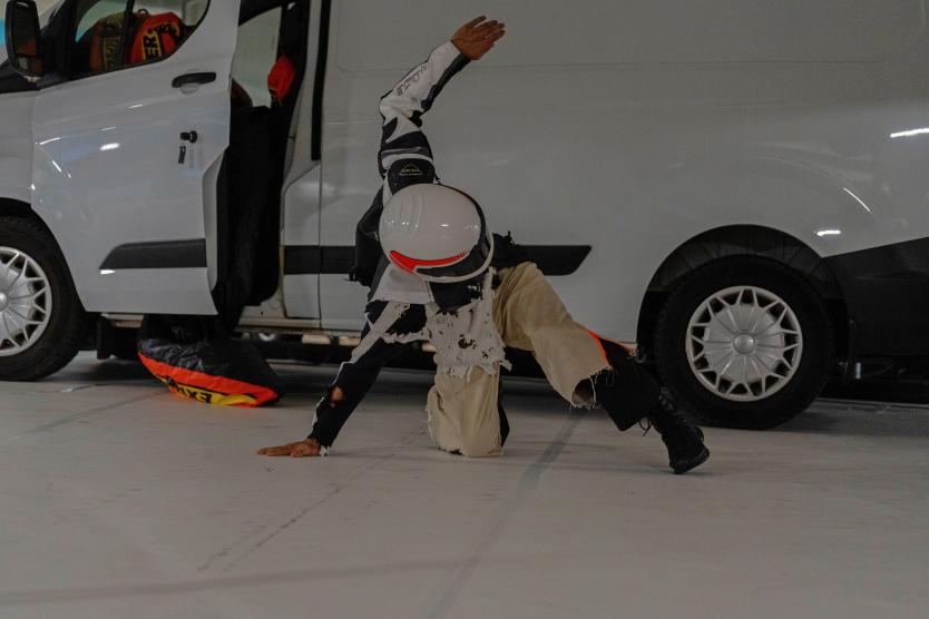 Ein Performer mit Motorradhelm und versetzter, dynamischer Kleidung stützt sich mit einer Hand auf den Boden vor einem weißen Lieferwagen mit geöffneter Seitentür, während der andere Arm gestreckt in die Luft zeigt.  A performer wearing a motorcycle helmet and layered, deconstructed clothing leans one hand on the floor in front of a white van with an open side door, extending the other arm upward in a dynamic pose. 