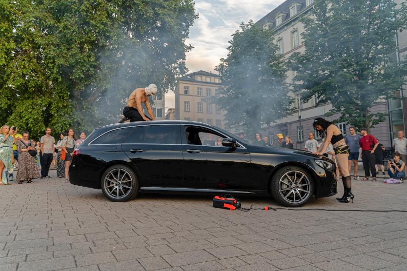 Eine Performance-Aktion im Freien um ein schwarzes Auto herum, auf dessen Dach eine Person kniet, umgeben von Publikum und leichtem Rauch.  An outdoor performance around a black car, with a person kneeling on its roof, surrounded by an audience and light smoke.