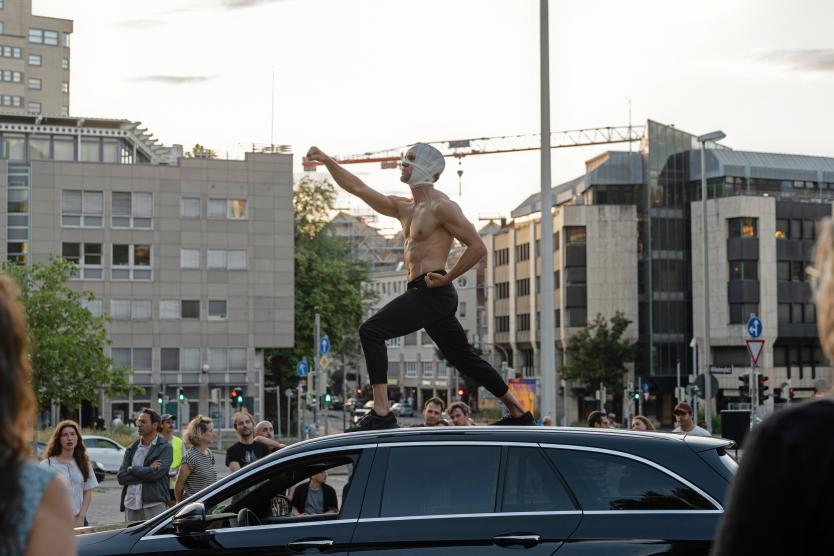 Ein Mann mit nacktem Oberkörper und bandagiertem Kopf steht in dynamischer Pose auf dem Dach eines schwarzen Autos vor einer städtischen Kulisse.  A bare-chested man with a bandaged head poses dynamically on the roof of a black car against an urban backdrop.