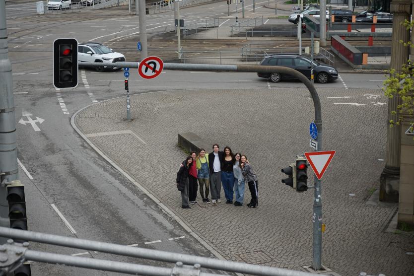 Aus der Vogelperspektive gesehen steht eine Gruppe junger Menschen eng zusammen auf einer gepflasterten Verkehrsinsel im städtischen Raum, umgeben von Ampeln und Straßen.   High-angle view of a group of young people standing closely together on a paved traffic island in an urban area, surrounded by traffic lights and roads. 