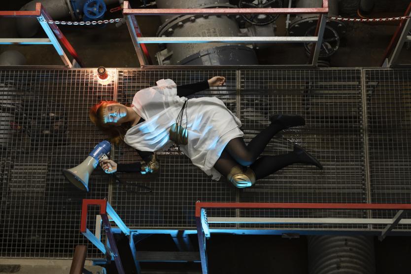 Vogelperspektive auf eine liegede rothaarige Person mit weißem Gewand. Die Umgebung ist eine dunkle Industriehalle, beleuchtet von blauem Licht. Die Person liegt auf einem Gittersteg und hält ein Megafon in der Hand.      Aerial view of a lying red-haired person in a white garment. The surroundings are a dark industrial hall illuminated by blue light. The person is lying on a metal grating walkway and holding a megaphone. 