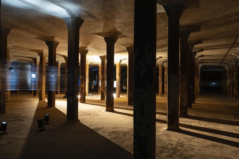 Betonsäulen in einem unterirdischen Raum. Der Raum wird von Baustrahlern ausgeleuchtet und wirkt wie eine Filmkulisse. Im Hintergrund ist zwischen zwei Säulen eine Folie gespannt. Concrete columns in an underground space. The room is illuminated by construction spotlights and looks like a film set. In the background, a sheet of plastic is stretched between two columns. 