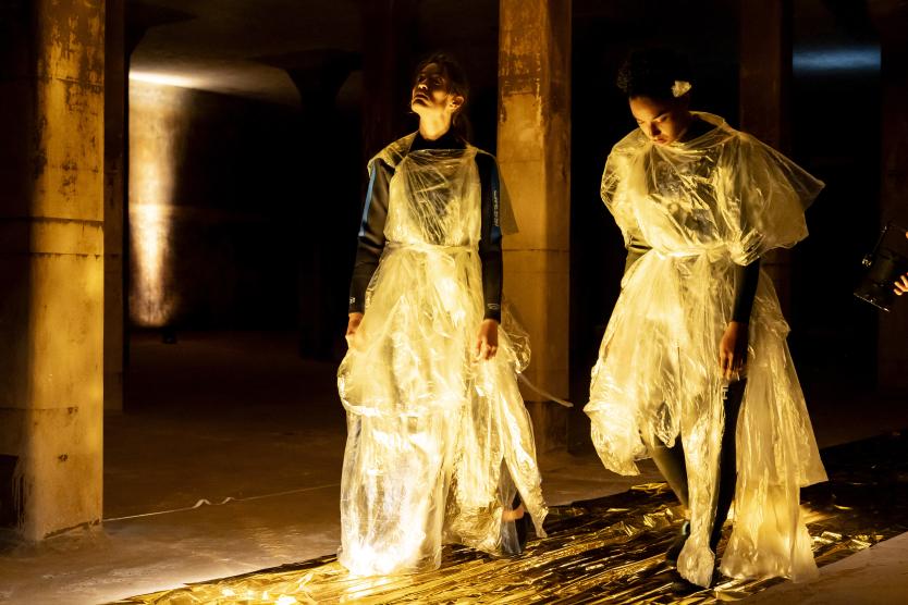 Zwei Performerinnen in Neoprenanzügen und durchsichtigen Plastikgewändern stehen in einem abgedunkelten, säulenbestandenen Raum auf einer gold glänzenden Folie. Two female performers wearing wetsuits and transparent plastic garments stand in a darkened, column-filled room on a shiny gold-coloured sheet of plastic. 