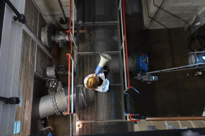 Draufsicht auf ein technisches Rohrsystem und Gitterroste in einem Industriegebäude, eine Person steht mit einem Megafon in der Mitte.      View from above of a technical pipe system and metal grating in an industrial building, with a person standing in the centre holding a megaphone. 