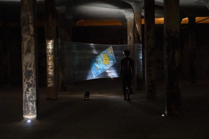 Silhouette einer Person in einem dunklen, mit Säulen durchzogenen Raum vor einer gespannten Folie, auf der eine bunte Karte abgebildet ist.  Silhouette of a person in a dark room lined with columns, standing in front of a stretched sheet of plastic displaying a colourful map. 
