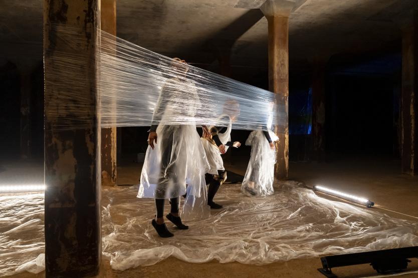Drei Performerinnen in Plastikkleidung bewegen sich hinter einem straff gespannten Band aus Klarsichtfolie zwischen Betonpfeilern. Three female performers in plastic clothing move behind a tightly stretched strip of transparent plastic sheeting between concrete pillars. 