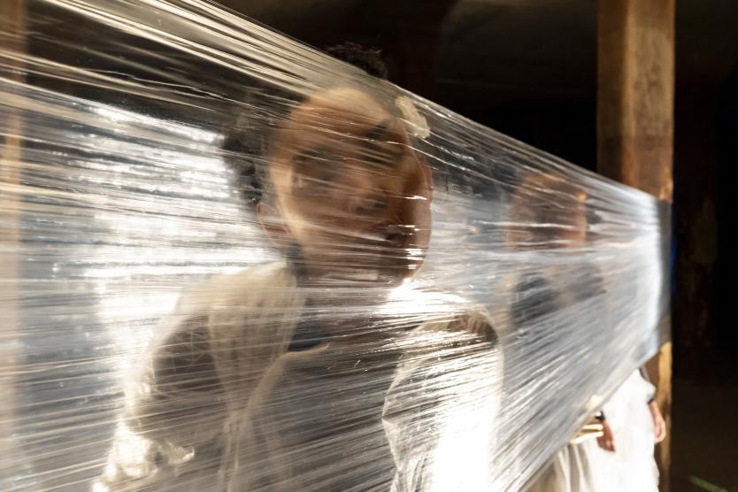Nahaufnahme des Gesichts einer Performering, das durch Schichten von gespannter, leicht durchsichtiger Folie gedrückt wird.  Close-up of a female performer's face pressed against layers of stretched, slightly transparent plastic sheeting. 
